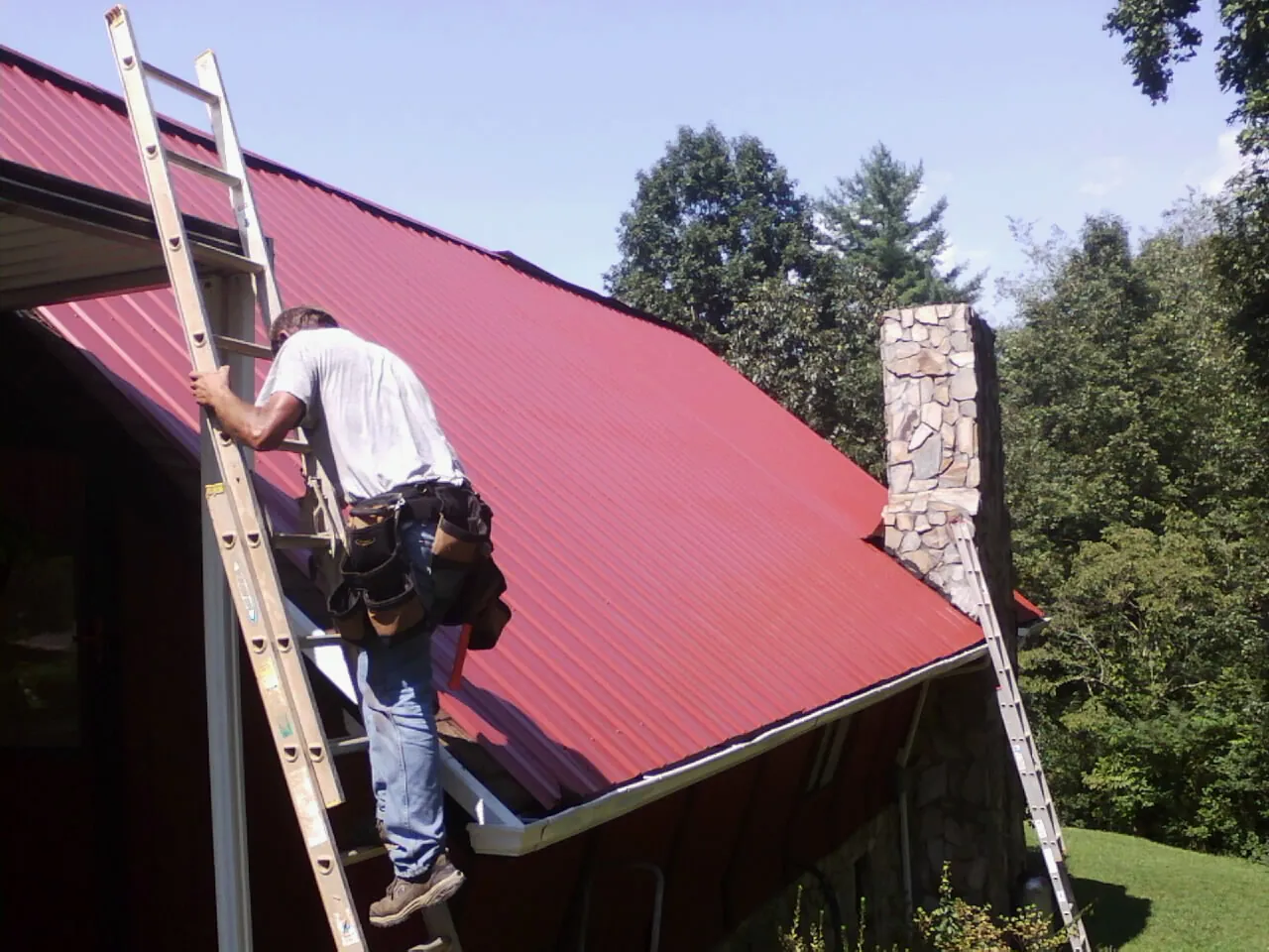 Roofer on a ladder beside a red metal roof and stone chimney — Soffit & Fascia Repair by Roofing Brothers in Jaffrey